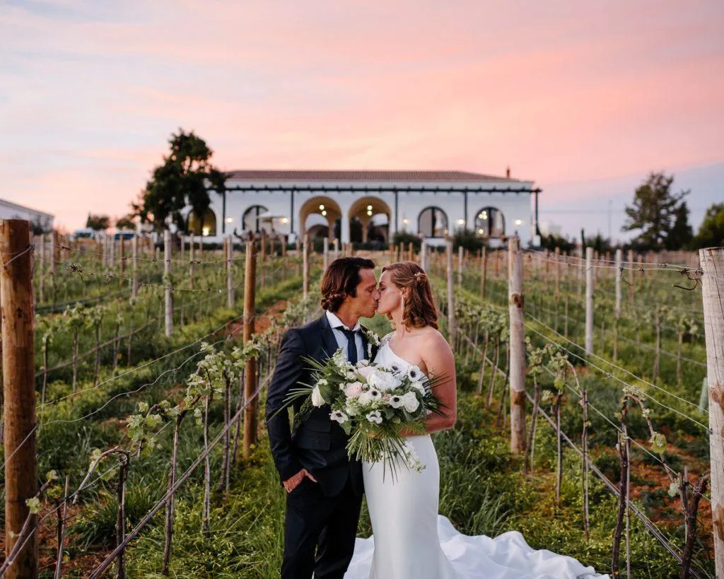 Couple kissing between rows of vines with a rose-coloured Algarve sunset behind them