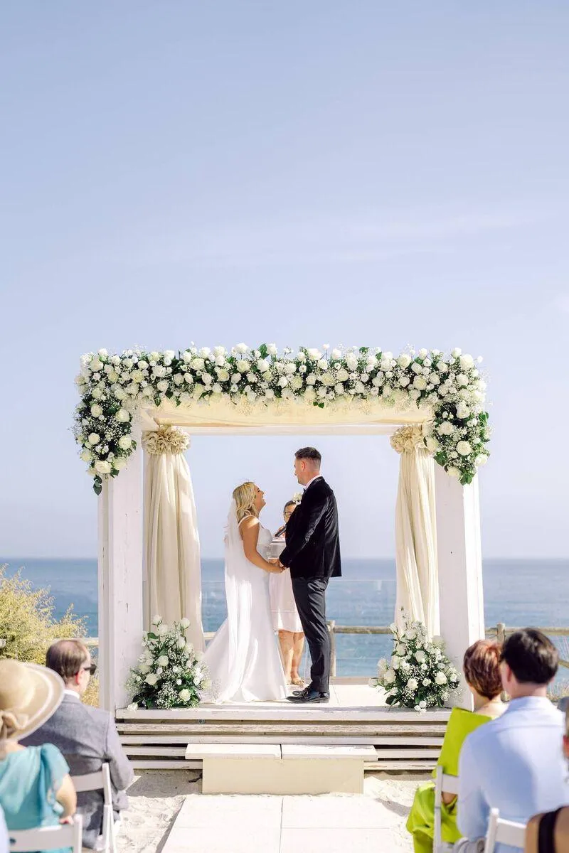 Bride and groom exchanging vows under a white floral chuppah above the sea