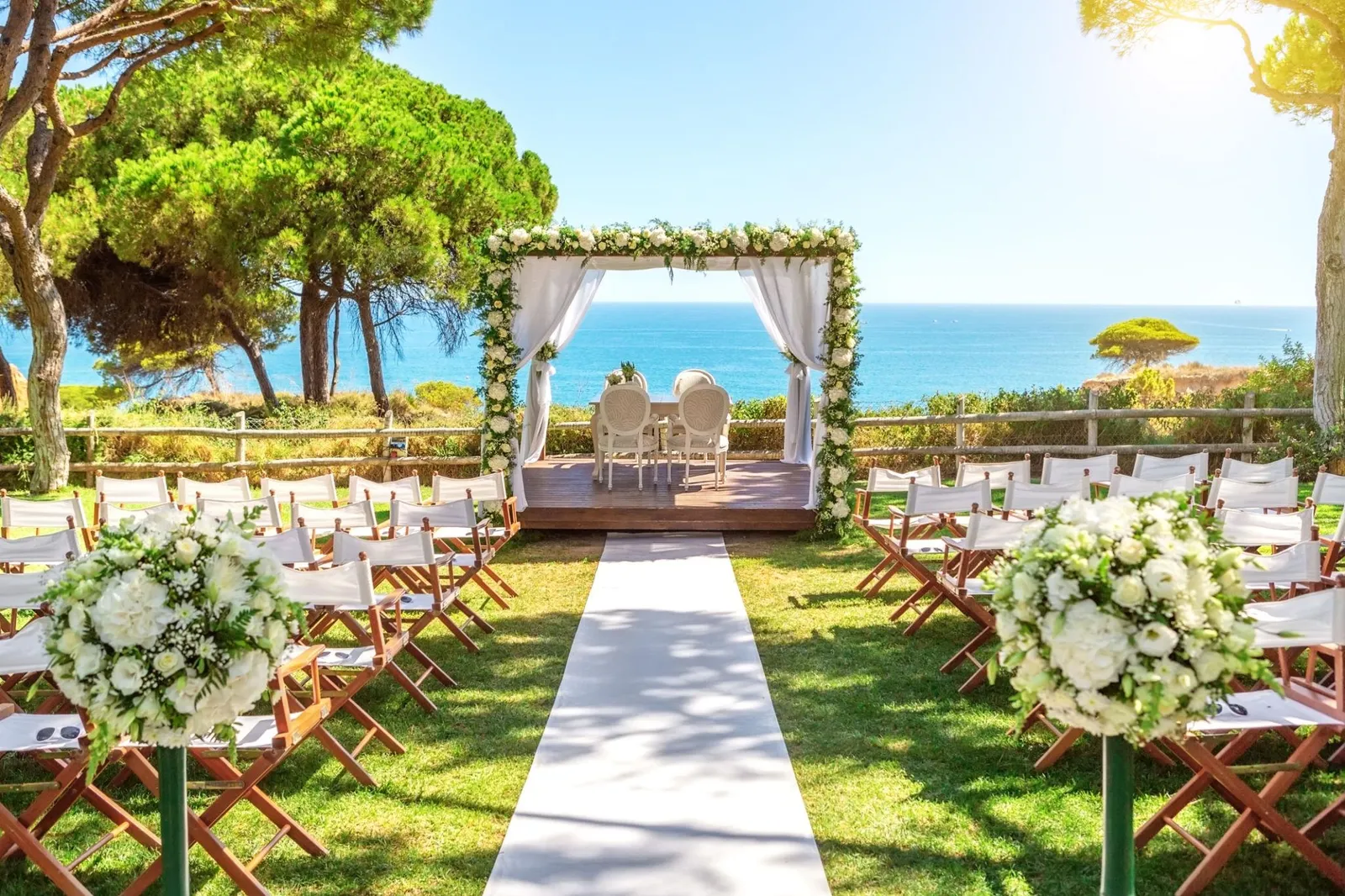 Sunlit chuppah dressed in white roses and greenery on a lawn above the sea