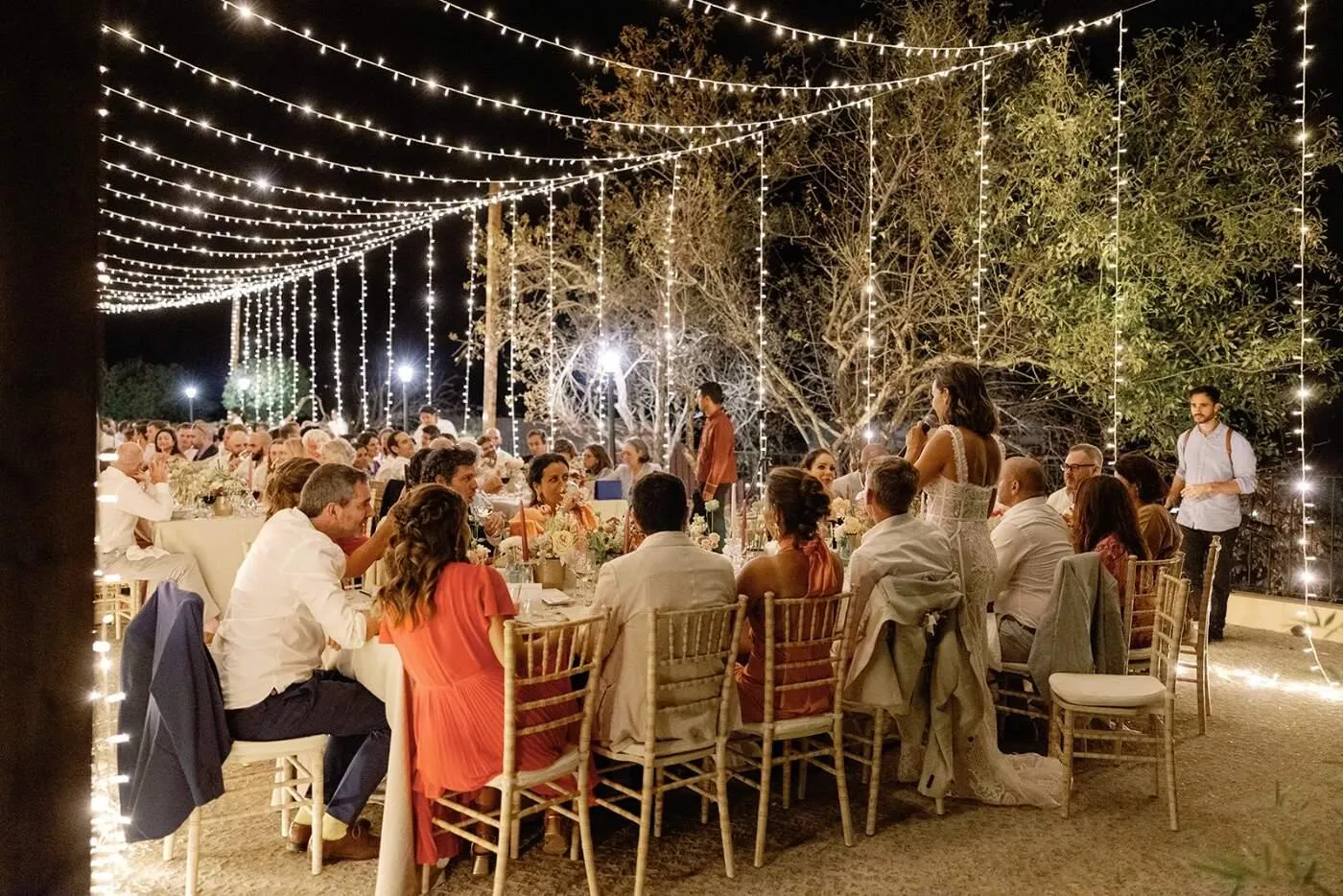 Guests gathered at a candlelit outdoor dinner beneath a suspended sky of fairy lights
