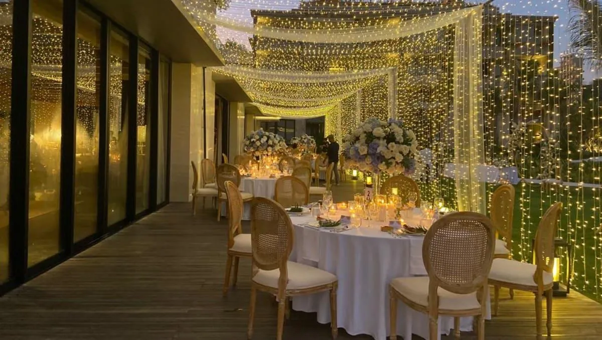 Terrace dinner table dressed in cane chairs beneath curtains of warm string lights